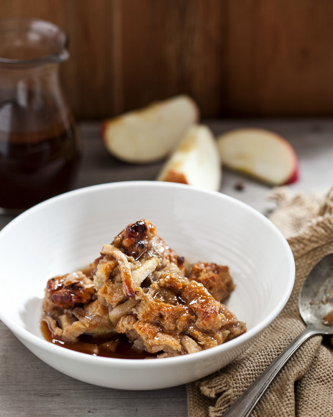 Baked apple bread pudding in a white bowl with apples and a jar of cinnamon-cider syrup in the background.